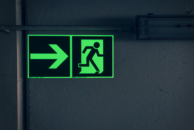 Cartel fluorescente de salida de emergencia, con una flecha señalando hacia la derecha y una figura humana saliendo por una puerta.